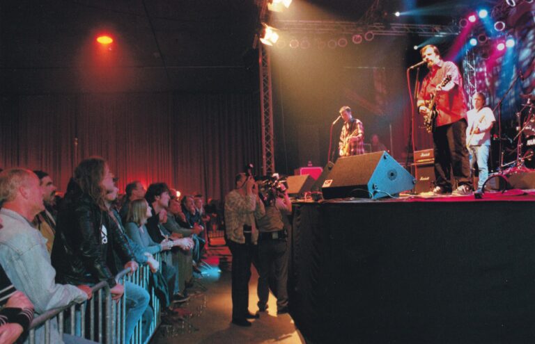 Een live bandoptreden met publiek dat naar het podium kijkt, verlicht door kleurrijke lampen.