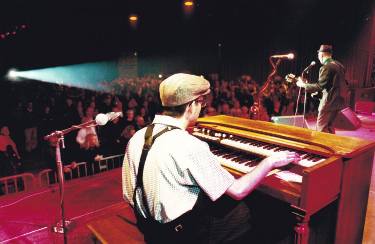 Man met een wit hemd en bretels achter een bruine piano op het podium.