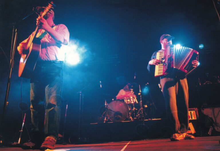 Drie muzikanten optreden op een podium met een drummer zichtbaar op de achtergrond, verlicht door kleurrijke lichten.