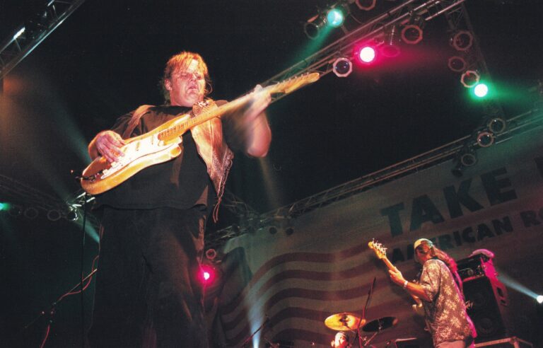 Een musician speelt gitaar op het podium tijdens een live optreden, met spots en lichteffecten op de achtergrond.