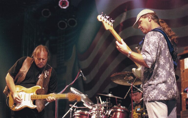 Twee muzikanten optreden op een podium met een drummer op de achtergrond.