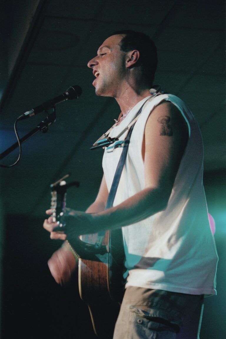 Man met een wit shirt aan die aan het optreden is met een gitaar en een mondharmonica