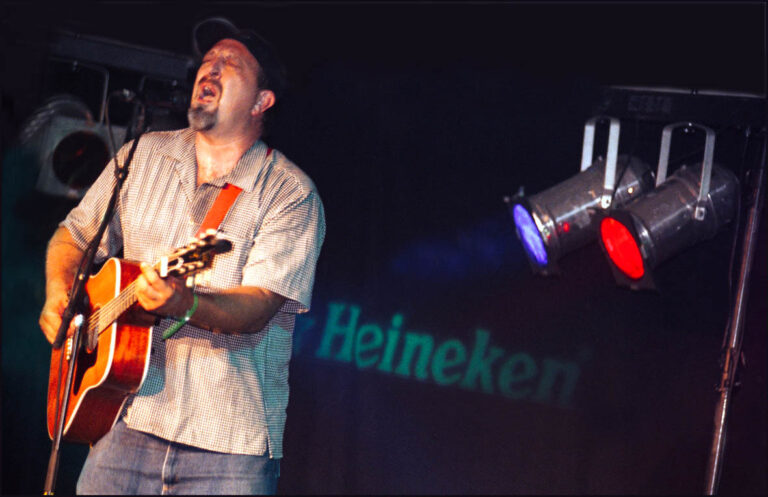 Man die aan het zingen is en de muziek begeleid met een gitaar. Achter hem zie je twee gekleurde lampen