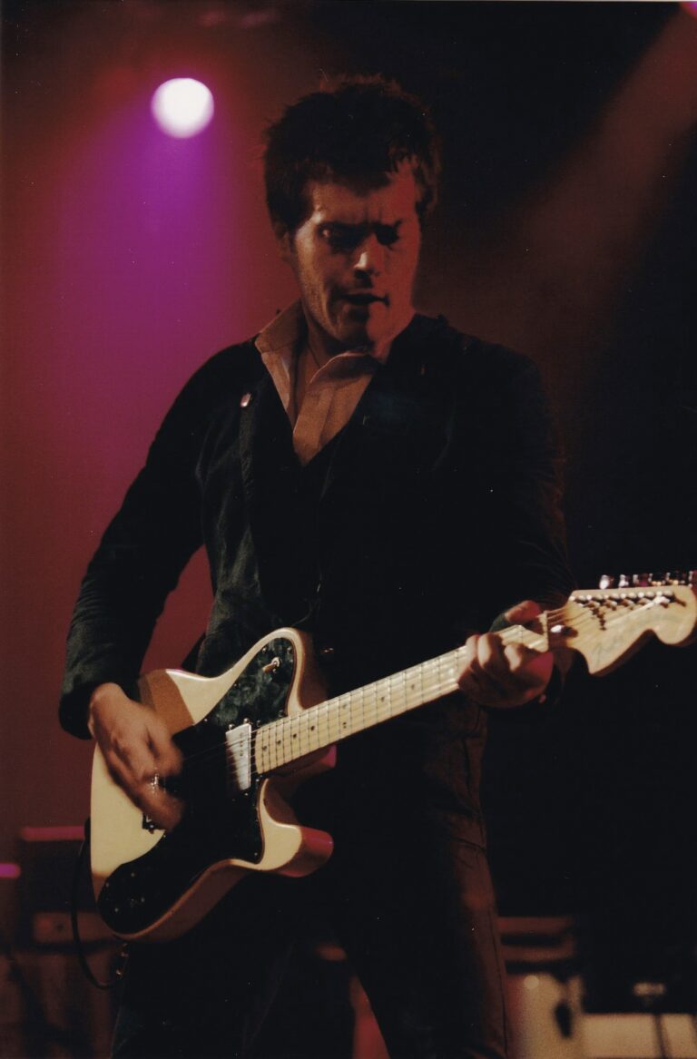 man met donker haar die aan het gitaar spelen. De achtergrond wordt fel verlicht door een roze lamp