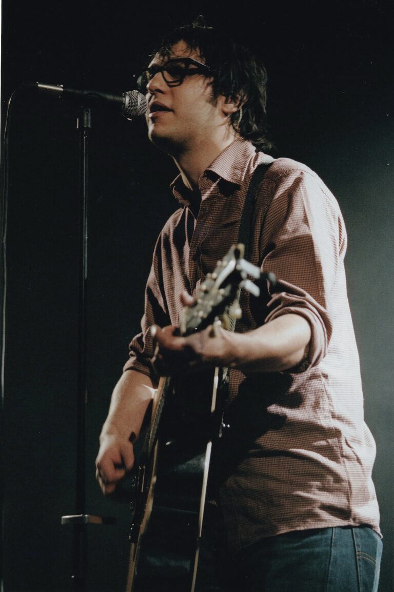 Man met een bril en donker haar die aan het gitaar spelen is terwijl hij aan het zingen is.