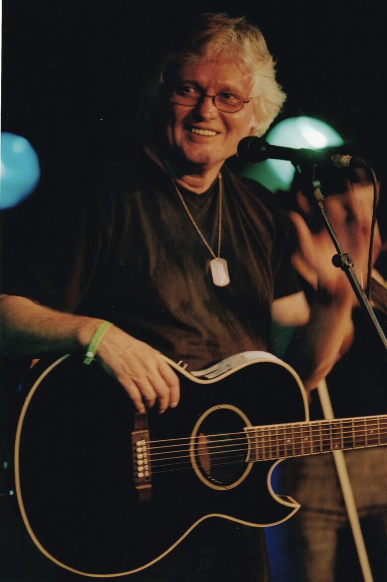 Een oudere man met een gitaar staat op een podium, terwijl hij glimlacht en zijn hand opsteekt.