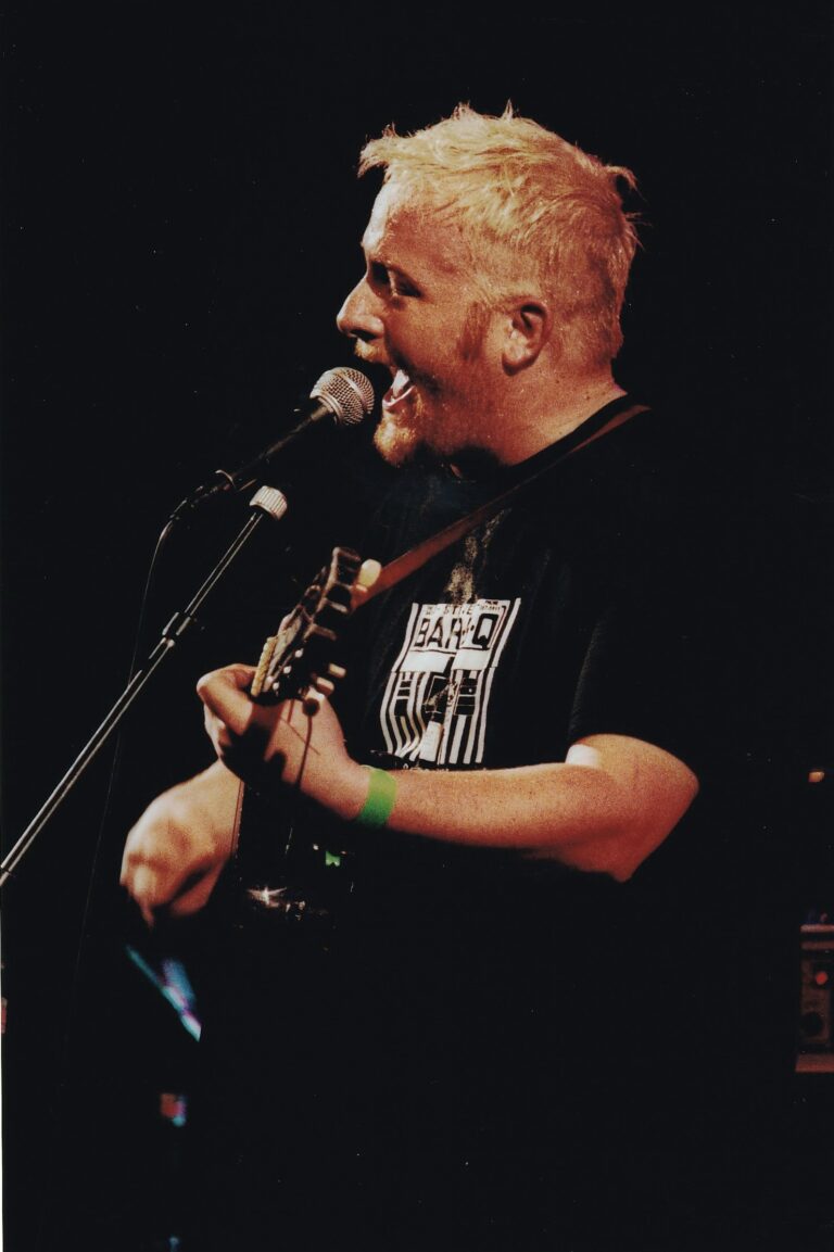 Een zanger met een gitaar staat op een podium en zingt in een microfoon.