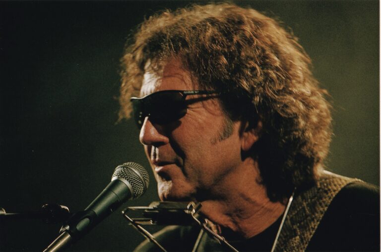 Man met krullend haar en een donkere zonnebril die dicht bij de microfoon gebogen staat.