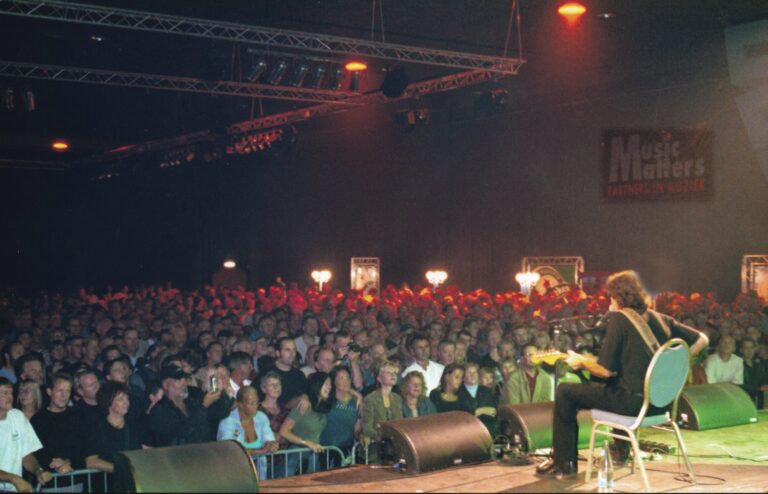 Een zanger of muzikant staat op een podium voor een groot publiek in een donkere hal, belicht door warme verlichting.