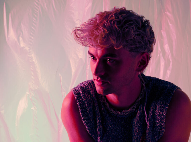 Een halfportret van een persoon met krullend haar, gekleed in een mouwloos shirt, met een kleurrijke achtergrond.