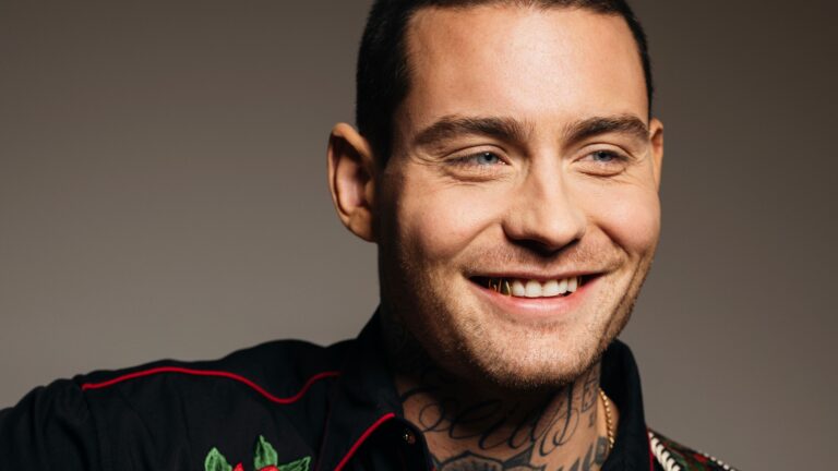 Een glimlachende man met een tatoeage op zijn hals, gekleed in een zwart shirt met kleurrijke borduursels.
