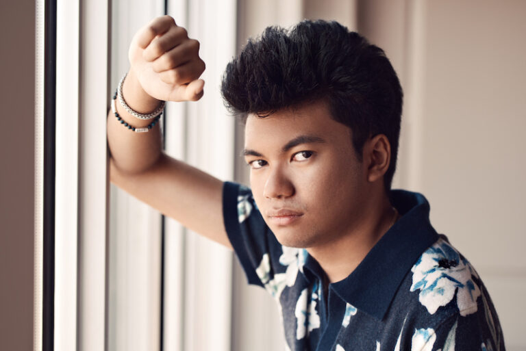 Een portretfoto van Joey Alexander waarbij hij nonchalant tegen een raam leunt en opzij de camera in kijkt.