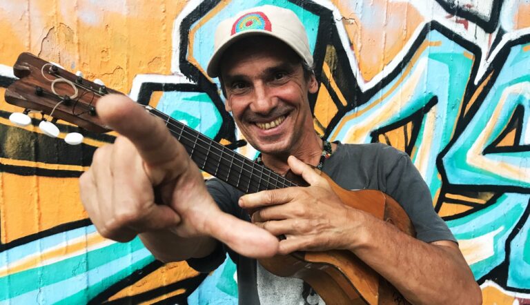 Manu Chao staat lachend voor een illustratie in graffiti-stijl met een gitaartje vinger wijzend