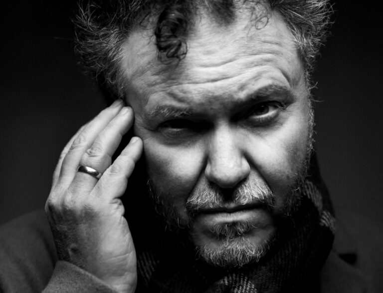 Een zwartwit portret van Jan Henk de Groot waarbij hij in de camera kijkt en zijn hand tegen de zijkant van zijn hoofd legt.