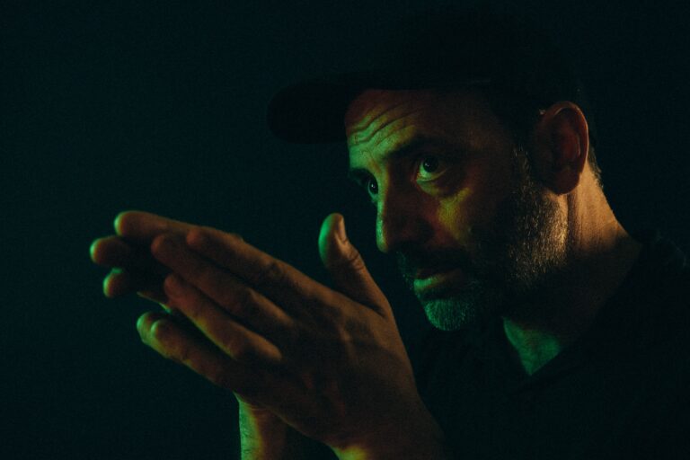 Persfoto van zanger Adrian Crowley die naar links kijkt an zijn handen tegen elkaar aan heeft.