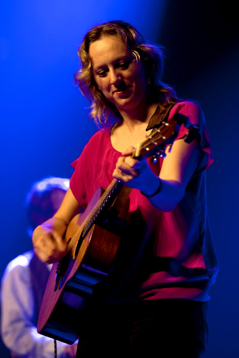 Een muzikante speelt gitaar op een podium, verblind door blauwe lichten.