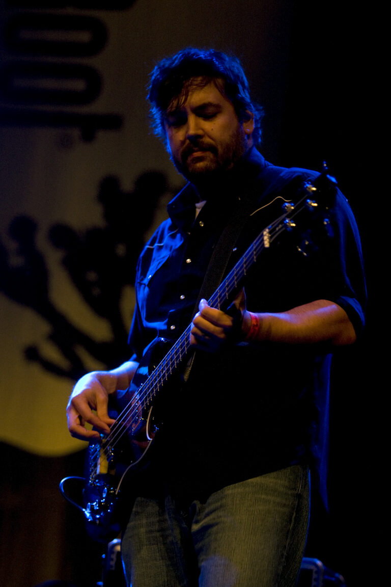 Een muzikant speelt basgitaar op een podium, omringd door een donkere achtergrond.