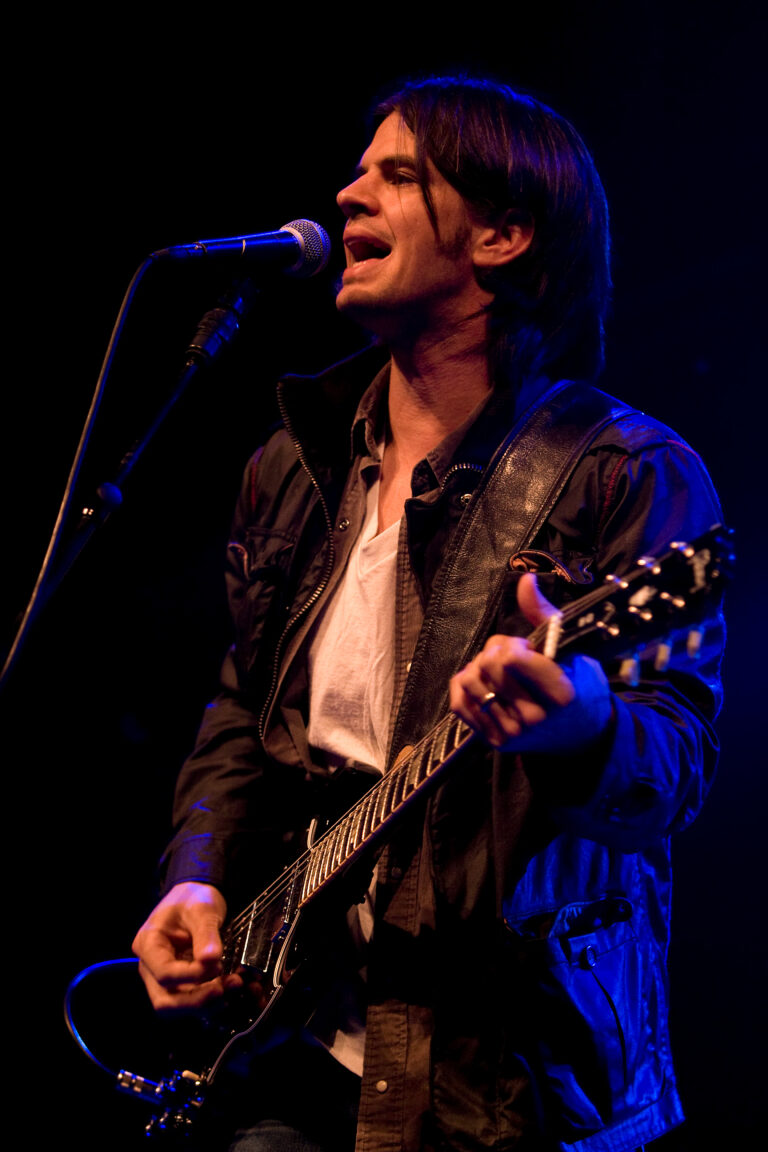 Een muzikant zingt en speelt gitaar op een podium met een microfoon in de hand.