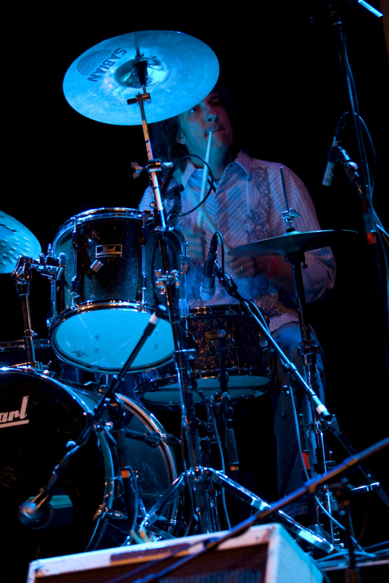 Een drummer bespeelt een drumstel tijdens een optreden in een donkere omgeving met blauwe verlichting.
