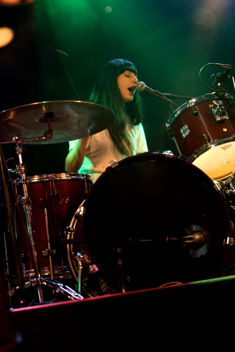 Een vrouwelijke drummer zingt en speelt op een drumstel in een sfeervolle, verlichte setting.