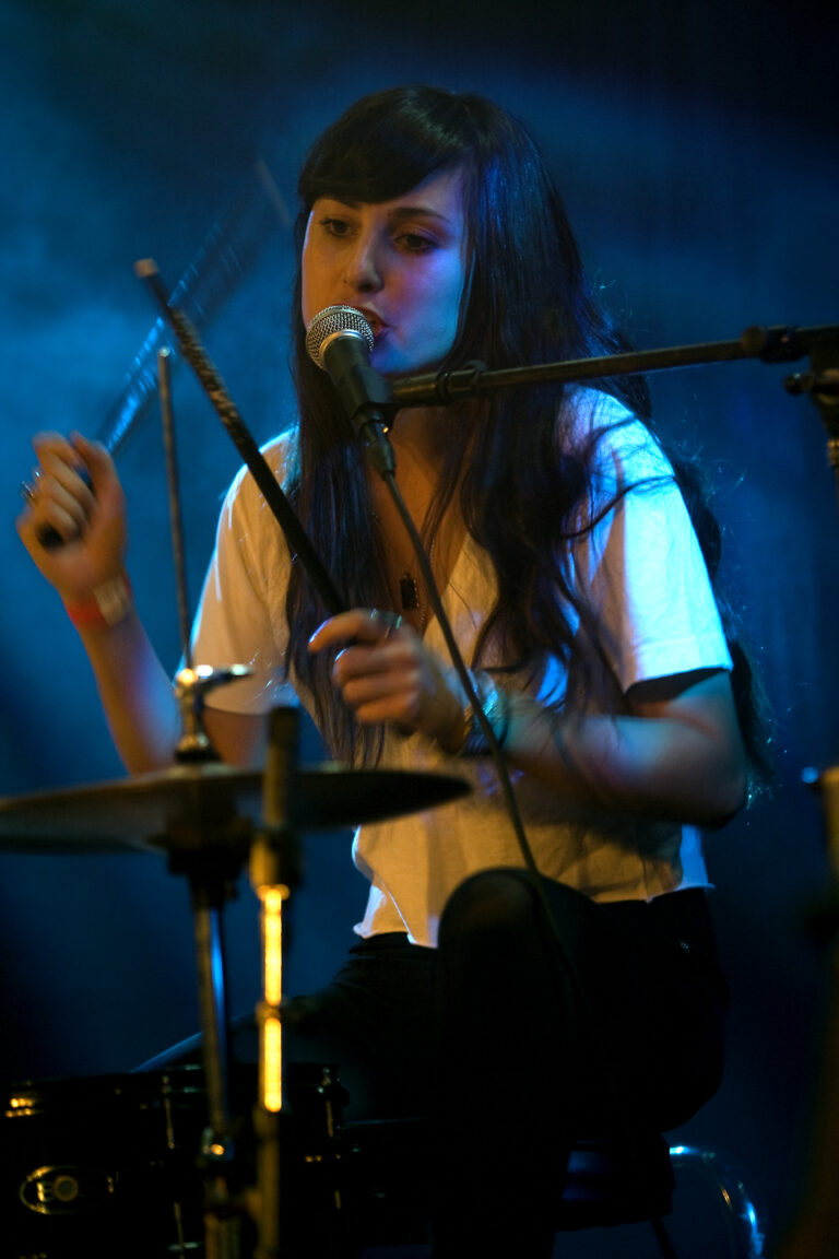Een jonge drummer speelt op een drumstel terwijl ze in een microfoon zingt, omringd door blauwe sfeerverlichting.