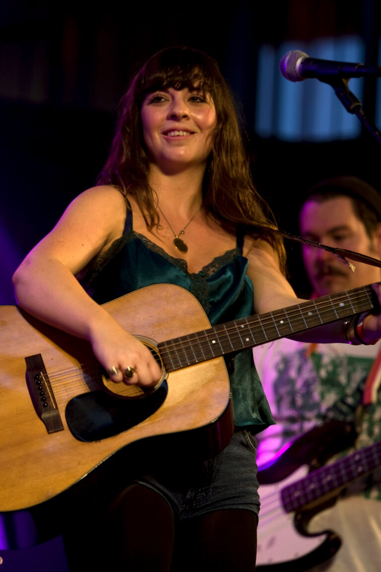 Een muzikant met een gitaar staat op het podium, terwijl ze zingt en naar het publiek kijkt.