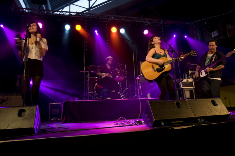 Een muzikale optredens op een podium met een zangeres die een gitaar speelt, terwijl een drummer op de achtergrond speelt, omringd door kleurrijke verlichting.