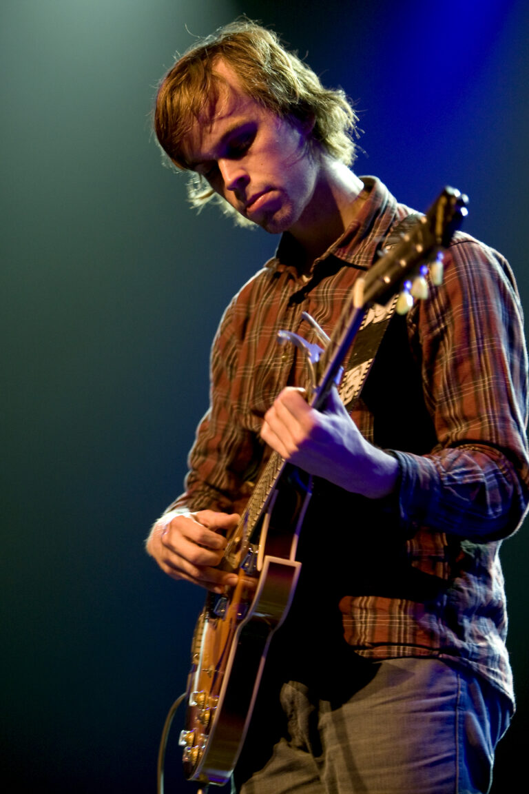 Een jonge muzikant speelt gitaar op een podium met een blauwe en paarse achtergrond.