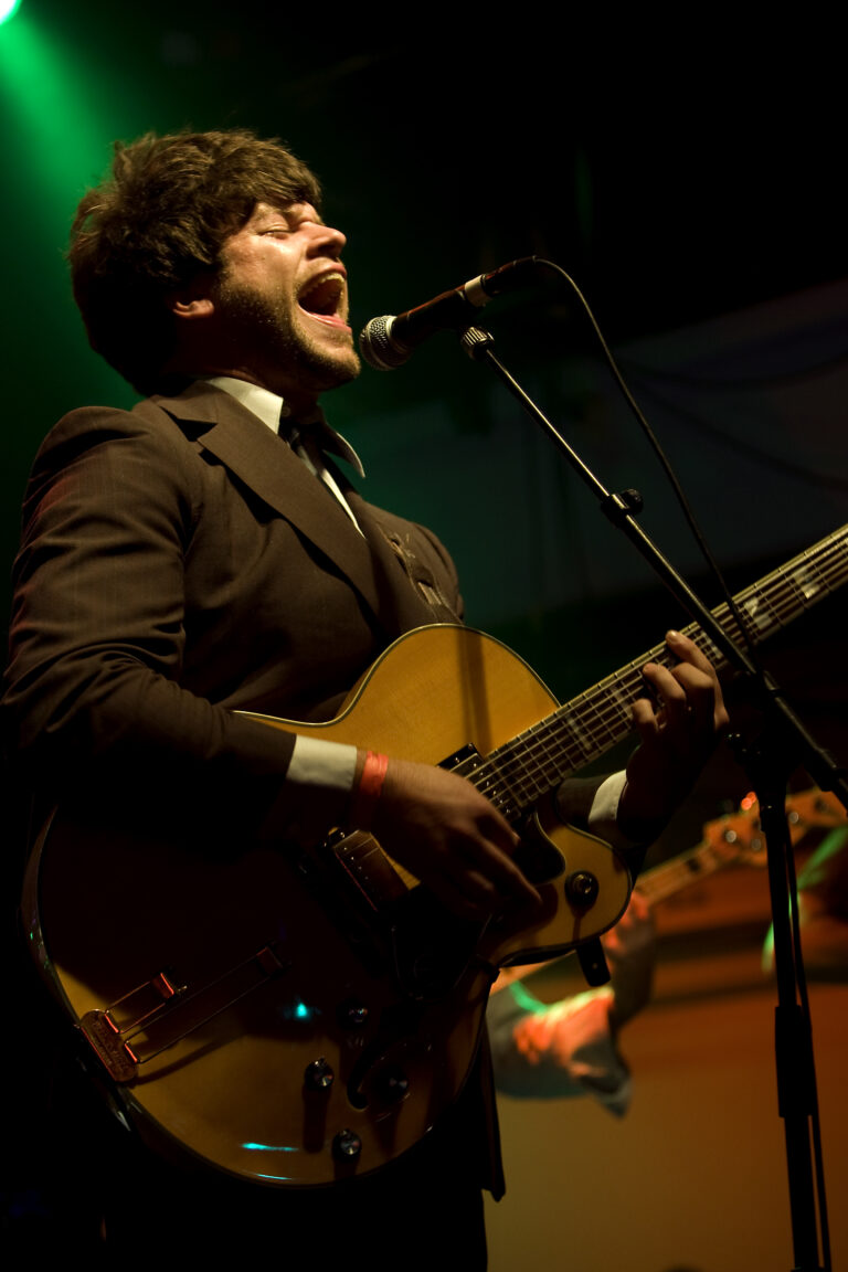 Een muzikant in een donkere outfit zingt en speelt gitaar op het podium, verlicht door groene lichten.