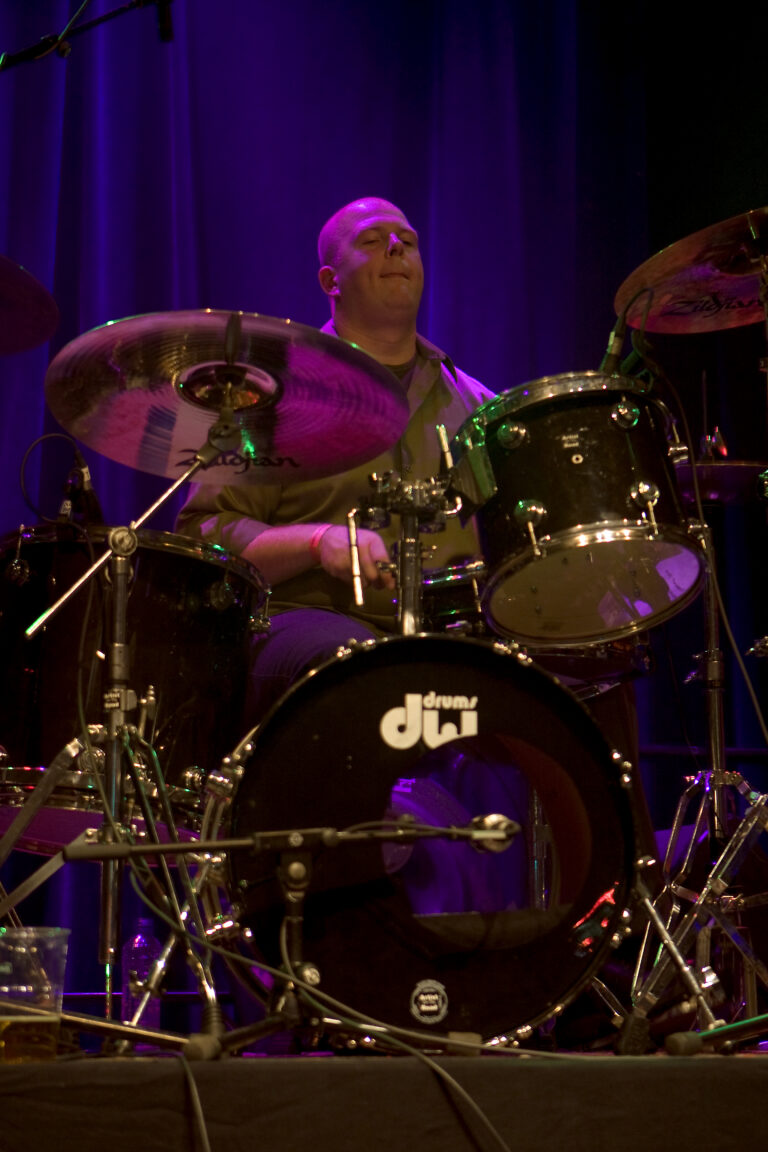 Een drummer speelt op een set drums, omgeven door paarse en blauwe achtergrondverlichting.
