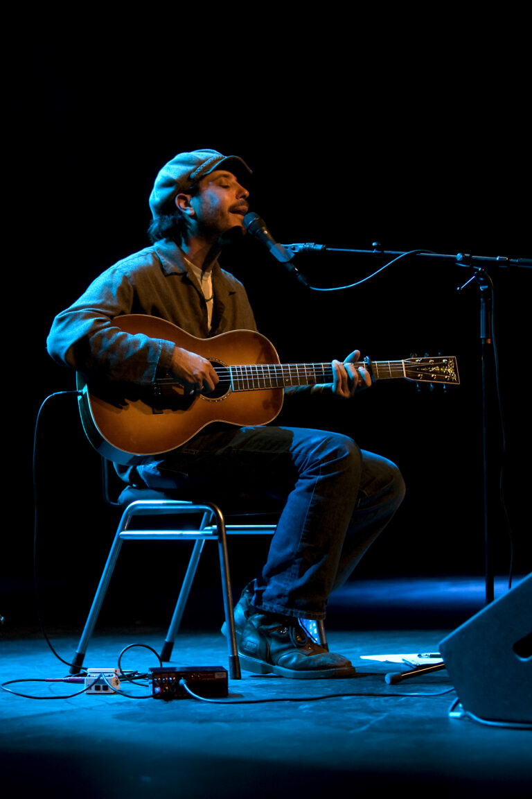 Een muzikant zit op een stoel en speelt gitaar terwijl hij zingt, verlicht door een blauwe gloed.