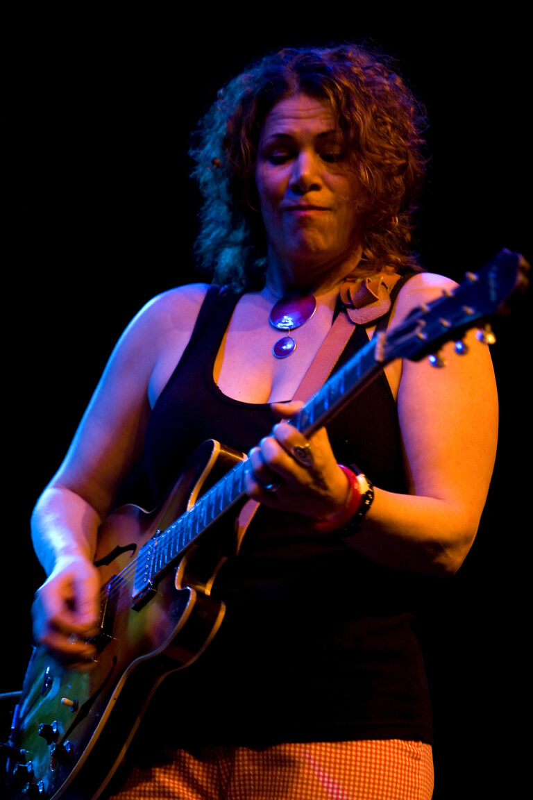 Een vrouw speelt gitaar op een podium met spannende verlichting.