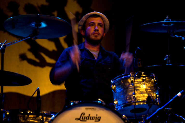 Een drummer in een hoed speelt op een drumstel met felgekleurde lichteffecten op de achtergrond.