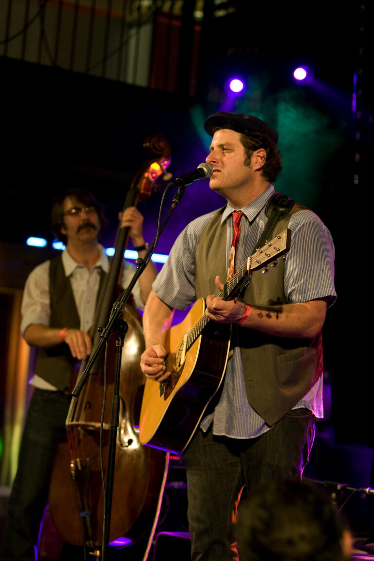 Twee muzikanten optreden op een podium, waarbij de voorste muzikant gitaar speelt en zingt.
