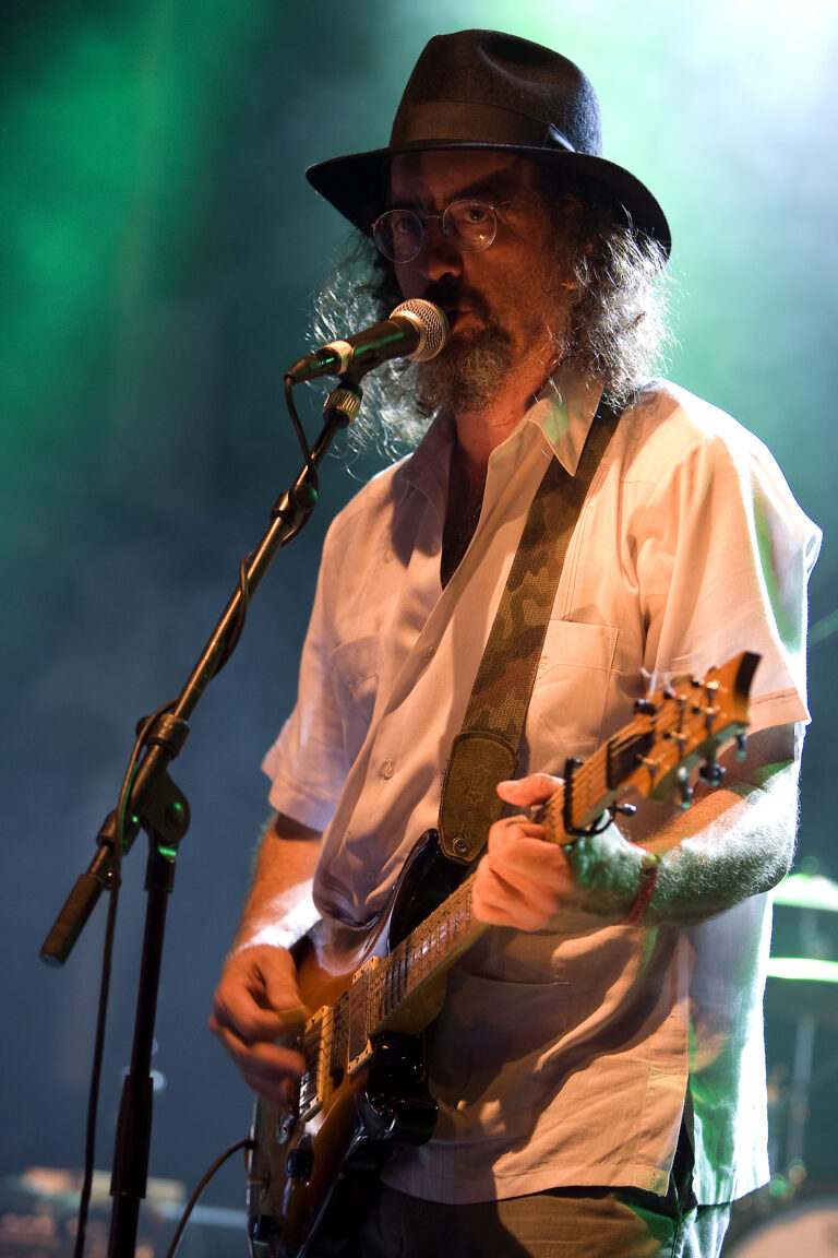 Een muzikant met een gitaar zingt in een microfoon op een podium met een groene achtergrond.