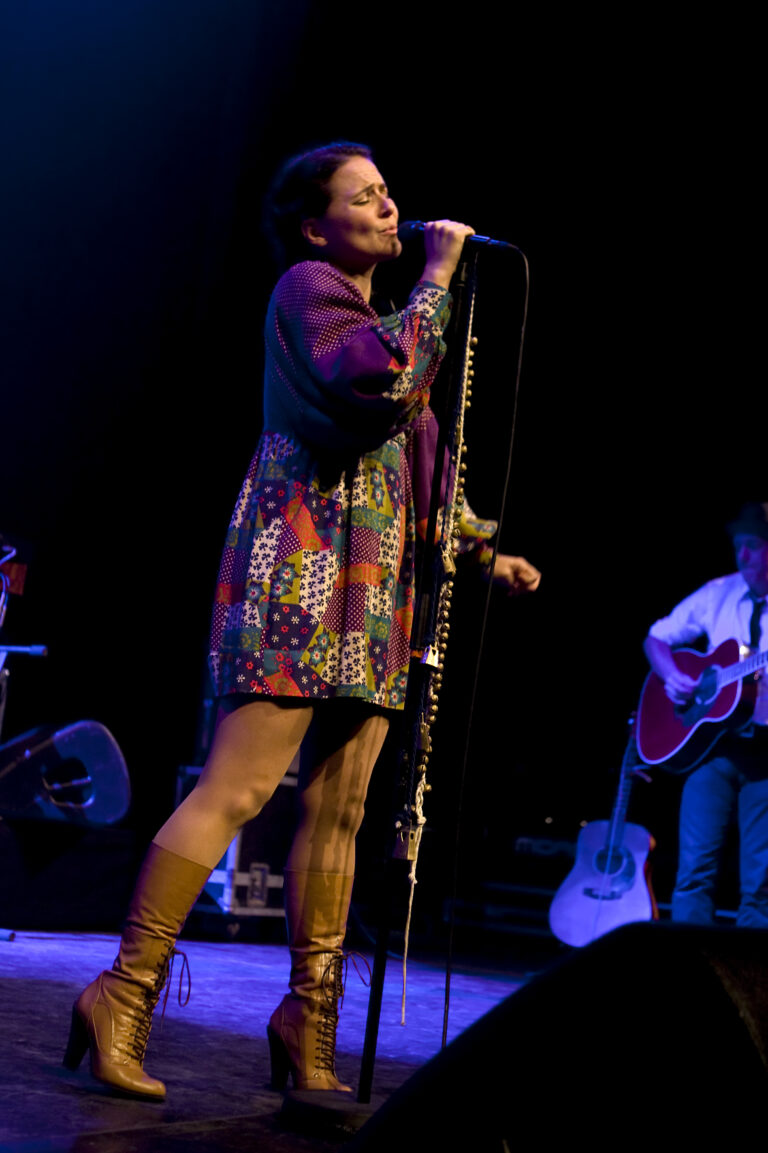 Een zangeres in een kleurrijke jurk treedt op met een microfoon, terwijl muzikanten op de achtergrond spelen.