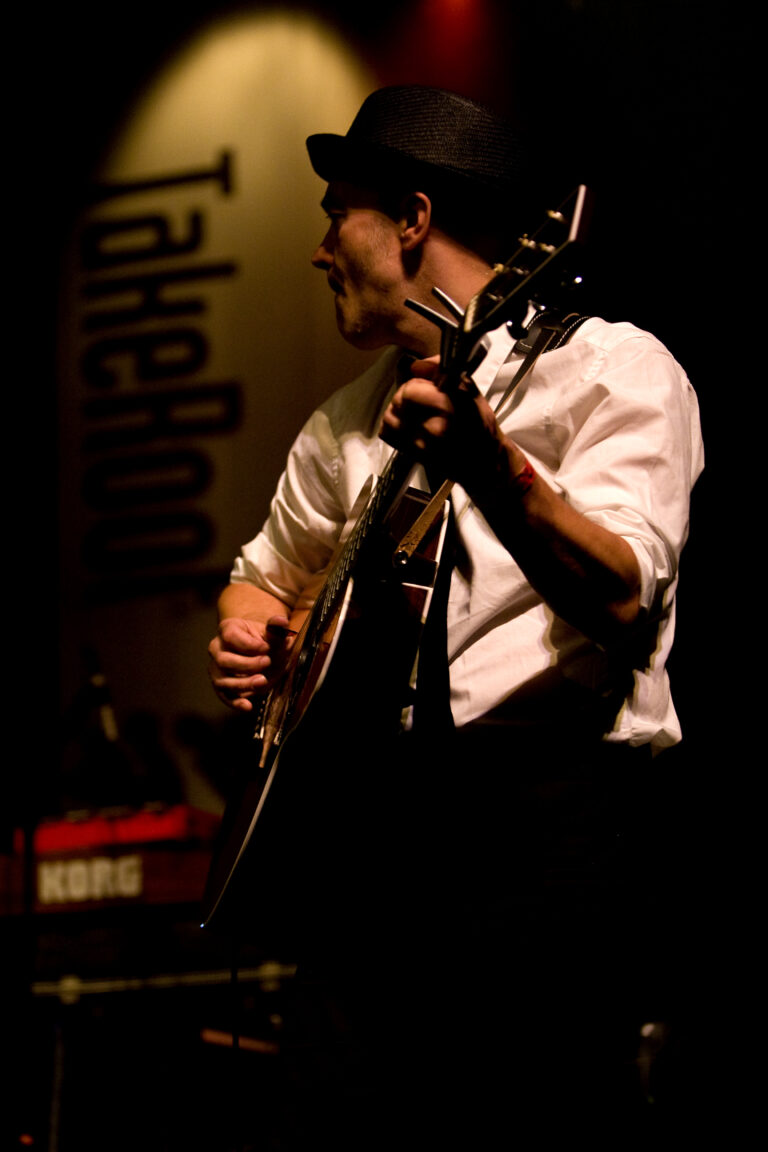 Een muzikant die een gitaar speelt op een podium, met een focus op zijn handen en het instrument in een sfeervolle, donkere setting.