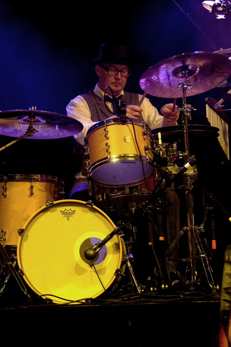 Een drummer speelt op een set met een gele basdrum en een paar cymbals, terwijl hij geconcentreerd is op zijn muziek.