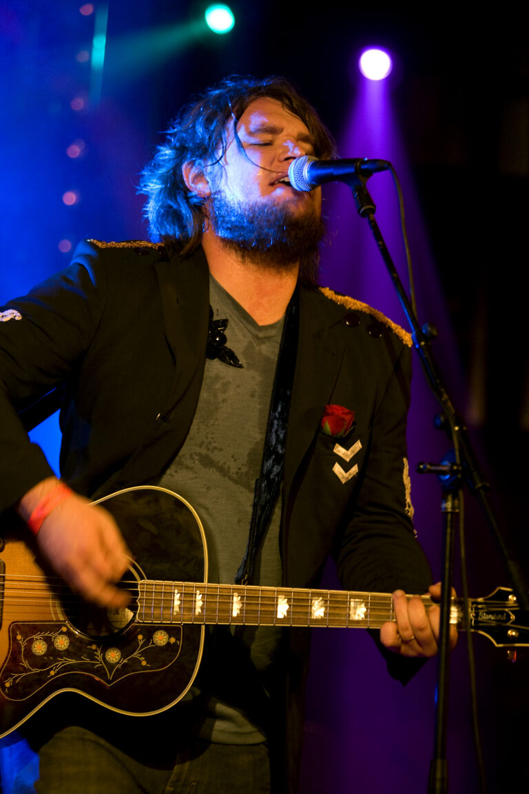 Een muzikant met een gitaar staat op een podium terwijl hij zingt, omringd door kleurrijk licht.