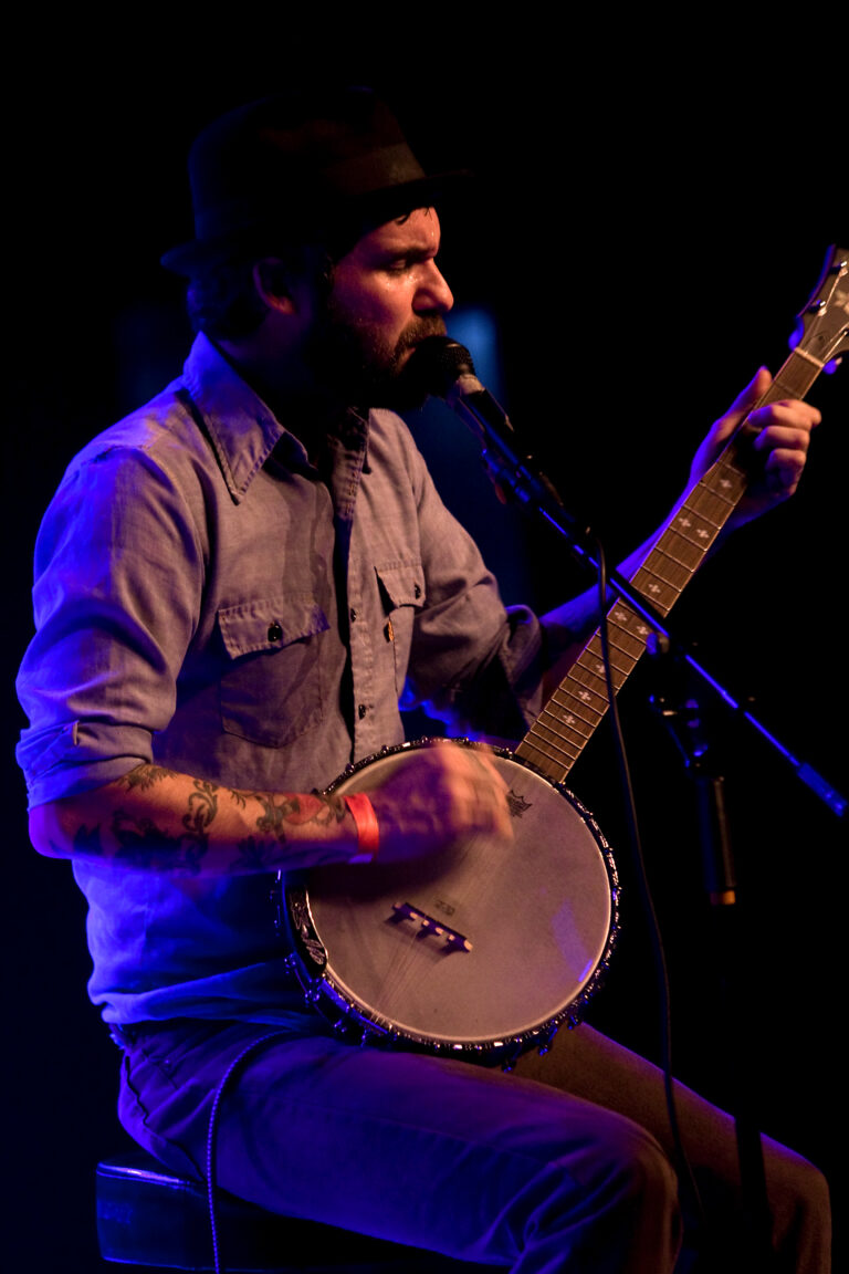 Een muzikant speelt op een banjo tijdens een optreden, omgeven door blauwe belichting.