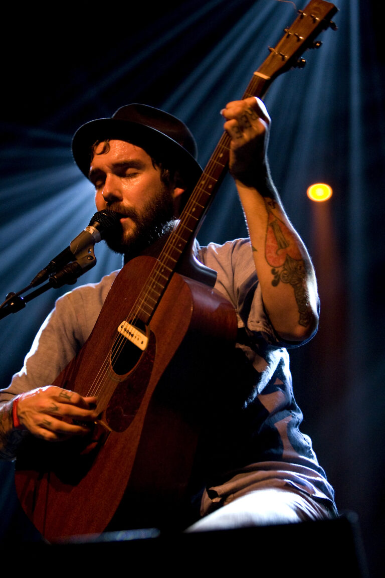 Een muzikant met een gitaar, zittend op een podium en omringd door dramatisch licht.