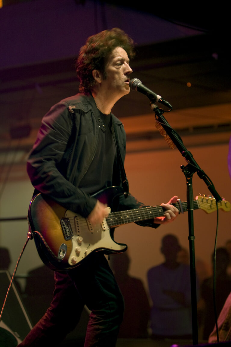 Een muzikant speelt gitaar en zingt op het podium, met een publiek op de achtergrond.