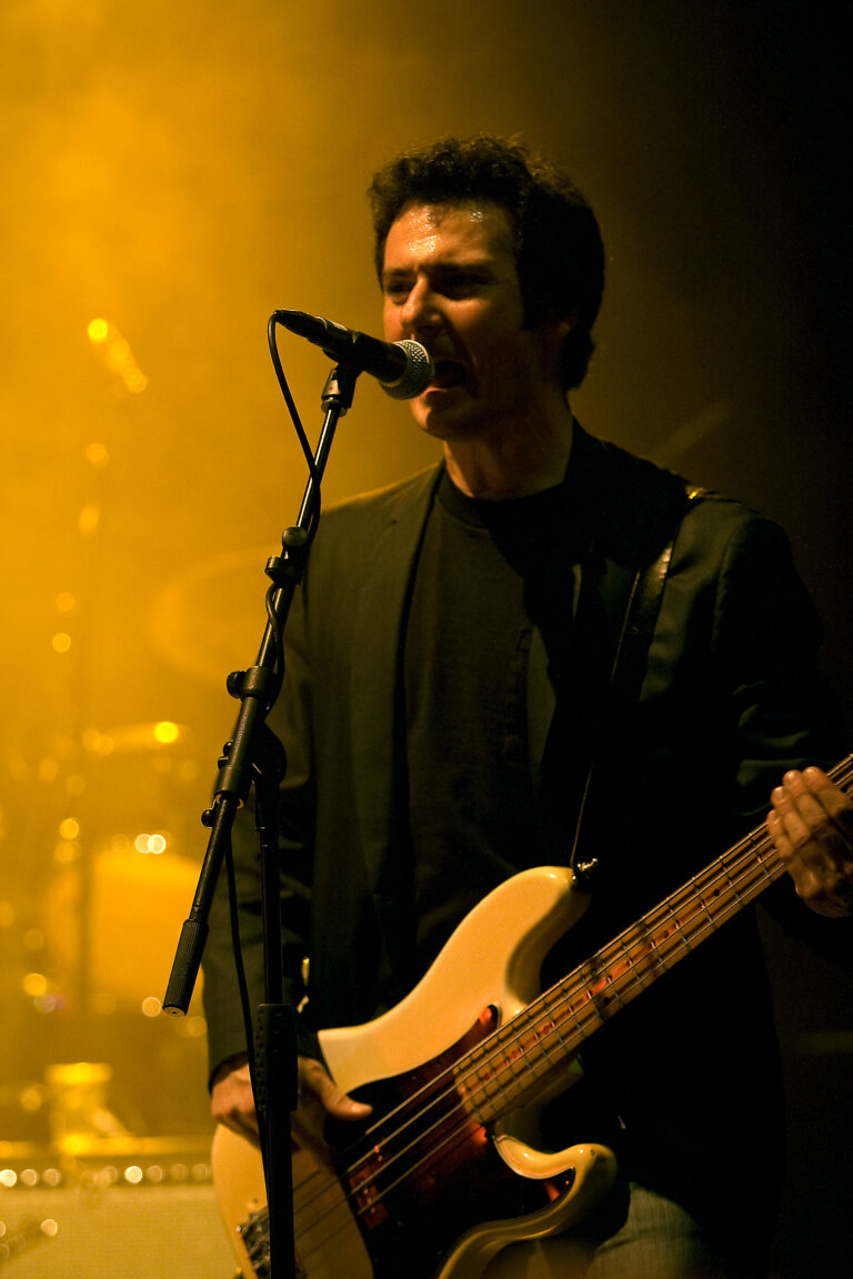 Een muzikant zingt en speelt basgitaar op een podium met een gele achtergrondverlichting.