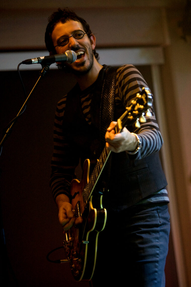 Een muzikant speelt gitaar en zingt in een microfoon, gekleed in een gestreept shirt en een vest.