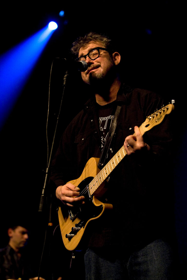 Een muzikant speelt gitaar en zingt op het podium, verlicht door een blauwe schijnwerper.