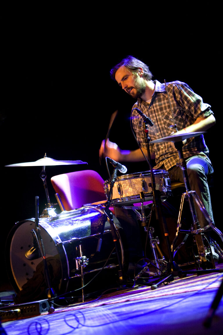 Een drummer in actie op het podium met een gitaar, terwijl hij stevig op de drums speelt.