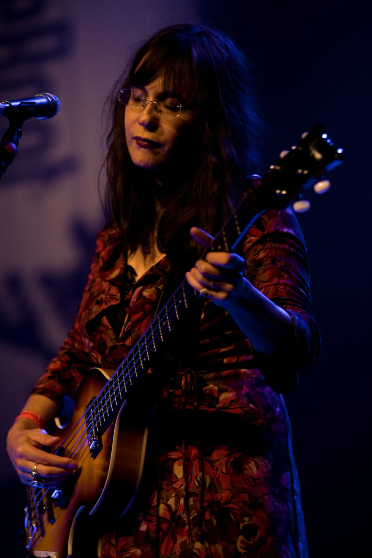 Een muzikante speelt gitaar op een podium met een blauwe achtergrond.