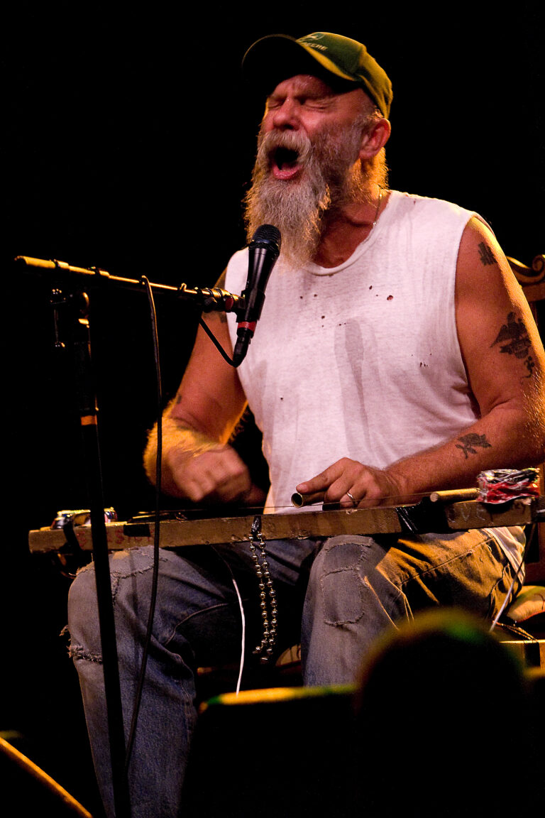 Een muzikant met een baard zingt en speelt op een zelfgemaakt instrument tijdens een optreden.