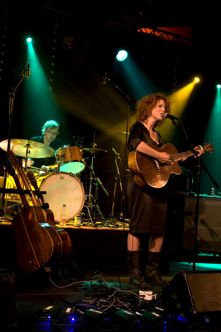 Een zangeres met een gitaar treedt op op een podium, terwijl een drummer achter haar speelt, omgeven door kleurrijke lichten.