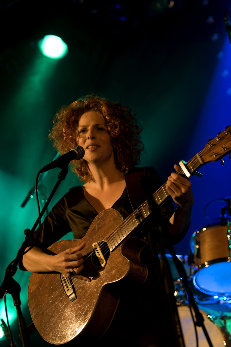 Een vrouw met krullend haar speelt gitaar en zingt op een podium met kleurrijke achtergrondverlichting.